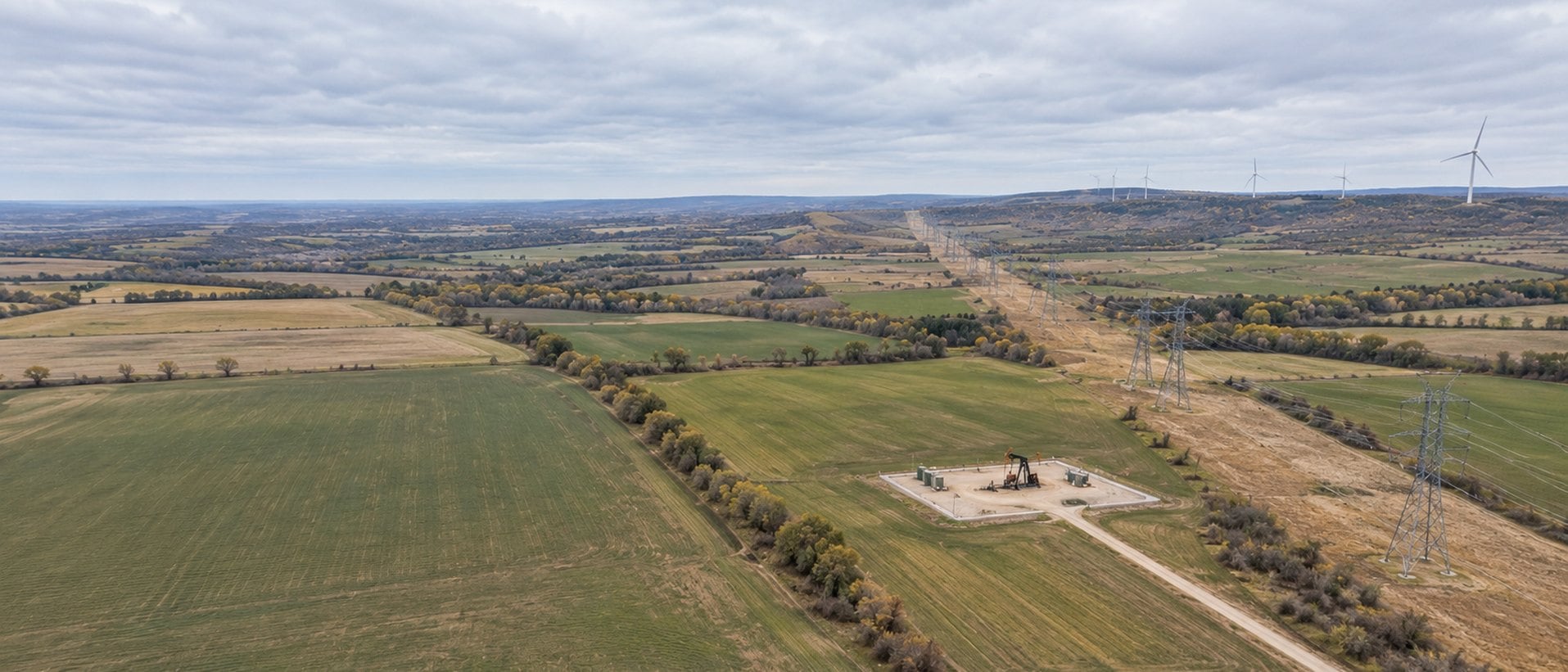 Drone view of farmland crossed by a power line right-of-way, with a pumpjack well pad and wind turbines beyond